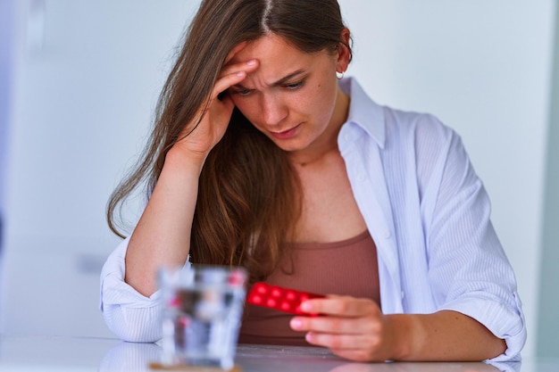 Unhealthy painful woman suffering from headache and holding blister pack with painkiller pills medicine at home Health problems