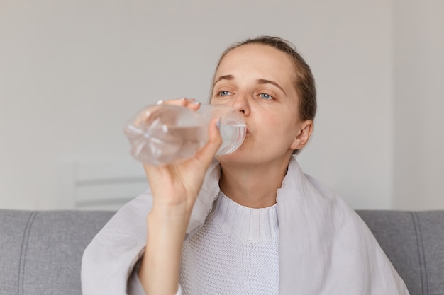 Unhealthy woman with hair bun sitting on sofa wrapped in blanket, having high temperature, drinking clear water to feel better, looking away with upset and tired expression.