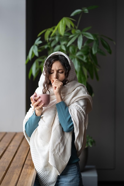 Unhealthy woman wrapped in plaid holding warm cup of tea and coughing feeling pain in throat