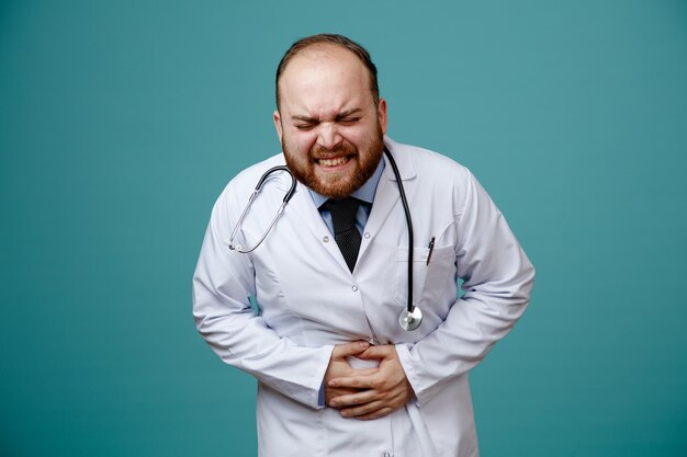 Unhealthy young male doctor wearing medical coat and stethoscope around his neck keeping hands on belly with closed eyes isolated on blue background