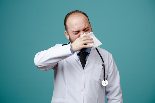 의료 코트와 청진기를 목에 두른 건강에 해로운 젊은 남성 의사는 파란색 배경에 격리된 눈을 감고 냅킨으로 코를 닦습니다