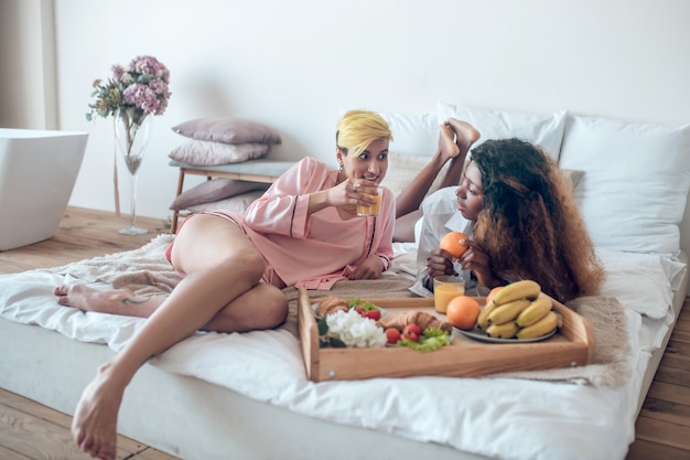 Unhurried morning. Couple of young adult women in sleeping clothes barefoot relaxed chatting on bed in morning