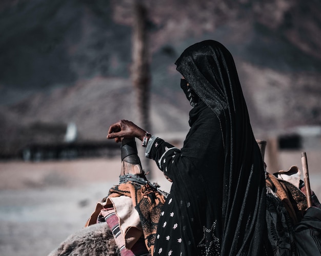 ラクダの鞍に手を置き、横たわるラクダの近くに立つ、アバヤを着た身元不明のベドウィン女性