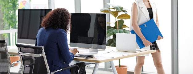 Unidentified businesswoman secretary officer staff in formal business suit sitting on chair working typing on computer PC at office table while other walking around.