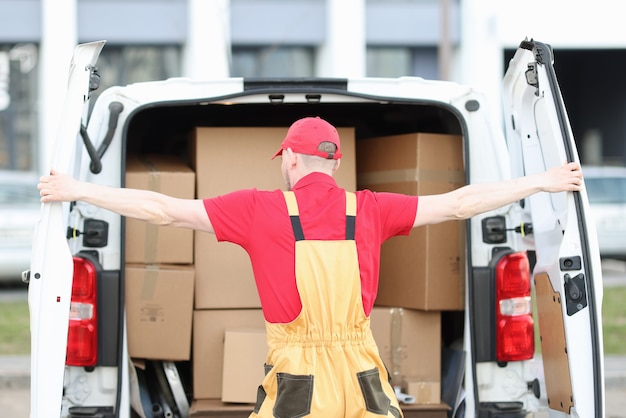 Il corriere maschio in uniforme apre il bagagliaio di un minivan pieno di scatole di cartone