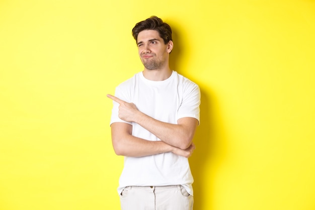 Unimpressed young man complaining, pointing and looking with skeptical face at bad product, standing against yellow wall