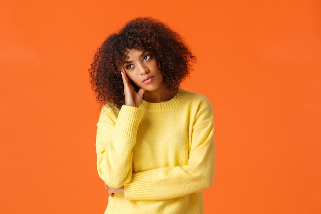 Uninterested bored african-american woman in yellow sweater, facepalm, looking away with uninterested, skeptical expression, attend boring party, standing upset and unsatisfied over orange 
