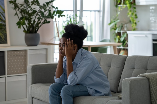 Uninterested discouraged African American woman sits on couch clutching head looking into distance