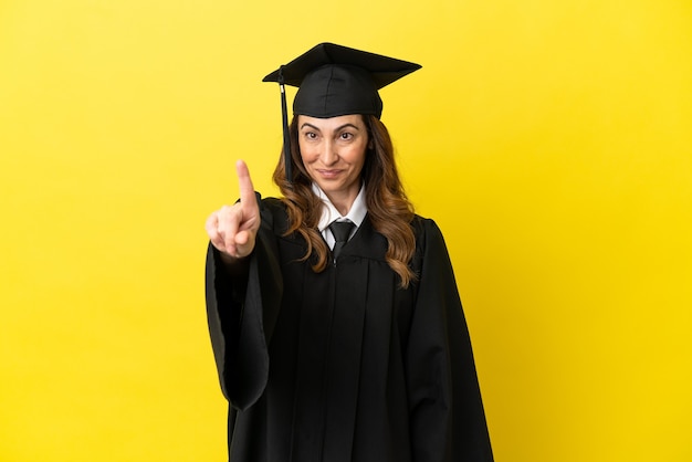 Universitair afgestudeerde van middelbare leeftijd geïsoleerd op een gele achtergrond die een vinger toont en optilt