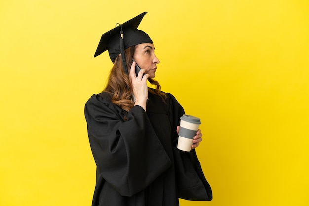 Universitair afgestudeerde van middelbare leeftijd geïsoleerd op gele achtergrond met koffie om mee te nemen en een mobiel