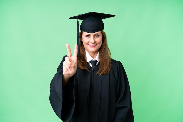 Universitair afgestudeerde vrouw van middelbare leeftijd over geïsoleerde achtergrond glimlachend en overwinningsteken tonend