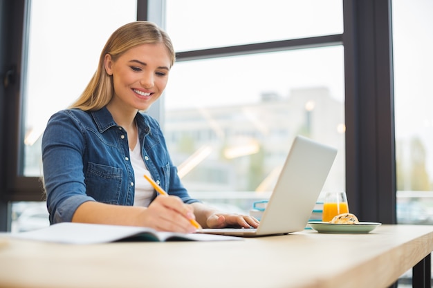 Universiteitsstudent. Vrolijke mooie positieve vrouw zittend in het café en bezig met de laptop terwijl ze haar huistaak doet