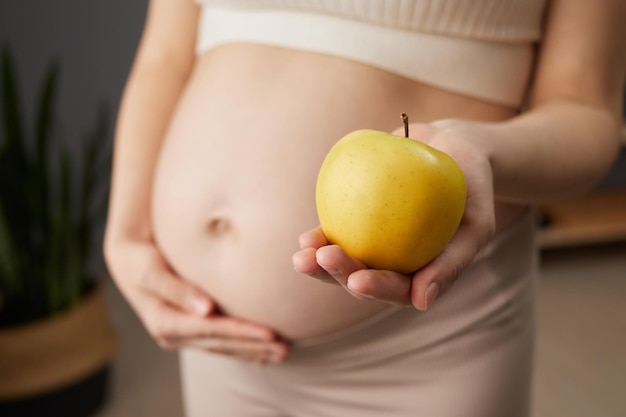 Фото Неизвестная красивая беременная женщина с голым животом стоит в домашнем интерьере с свежим органическим желтым яблоком предпочитает здоровое питание во время ожидания ребенка