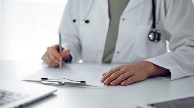 Unknown doctor woman sitting and writing notes at the desk in clinic or hospital office, close up. Medicine concept.