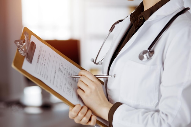 Unknown female doctor using clipboard in sunny clinic. Perfect medical service in hospital.