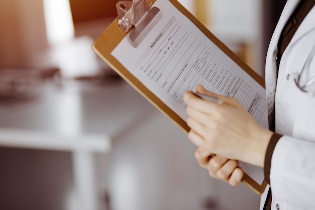 Unknown female doctor using clipboard in sunny clinic. Perfect medical service in hospital.