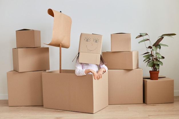 Photo unknown girl sitting surrounded with packages with personal pile, making head from cardboard box with drown smiling face, relocating, moving, real estate.