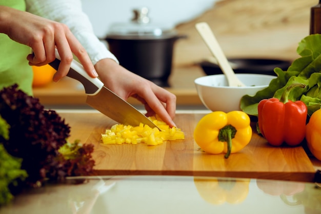 Unknown human hands cooking in kitchen. Woman slicing yellow bell pepper. Healthy meal, and vegetarian food concept.