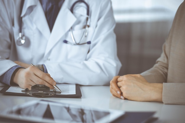 Unknown male doctor and patient woman discussing something while sitting in clinic and using clipboard. Best medical service in hospital, medicine, pandemic stop