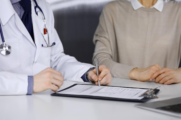 Unknown male doctor and patient woman discussing something while sitting in clinic and using clipboard. Best medical service in hospital, medicine, pandemic stop.