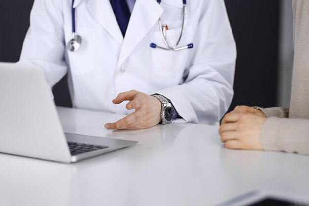 무명의 남성 의사와 환자 여성이 클리닉에 앉아서 노트북을 사용하는 동안 무언가에 대해 논의하고 있습니다. 병원, 의학, 전염병 중지에서 최고의 의료 서비스.
