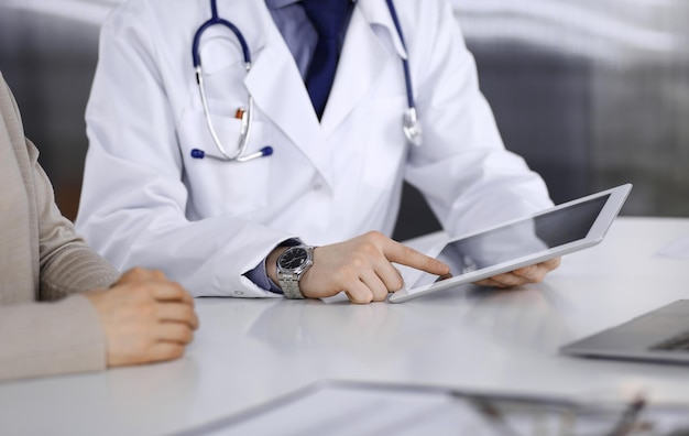 Unknown male doctor and patient woman discussing something while sitting in clinic and using tablet computer. Best medical service in hospital, medicine, pandemic stop.