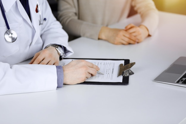 Unknown male doctor and patient woman discussing something while sittingin a darkened clinic and using clipboard, glare of light on the background.