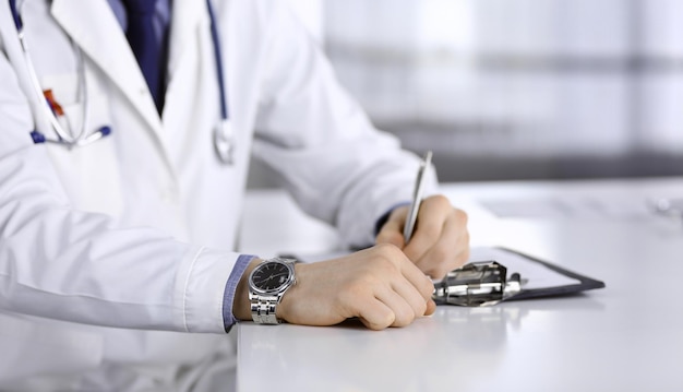 Unknown male doctor sitting and working with clipboard of medication history record in clinic at his working place, close-up. Young physician at work. Perfect medical service, medicine concept.