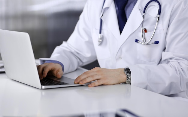 Unknown male doctor sitting and working with laptop in clinic\
at his working place, close-up. young physician at work. perfect\
medical service, medicine concept.