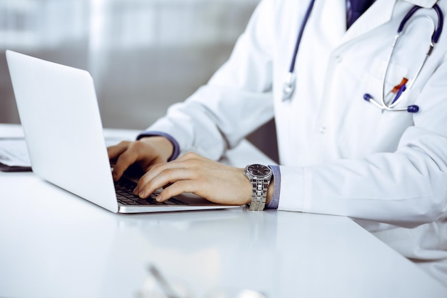 Unknown male doctor sitting and working with laptop in clinic at his working place, close-up. Young physician at work. Perfect medical service, medicine concept