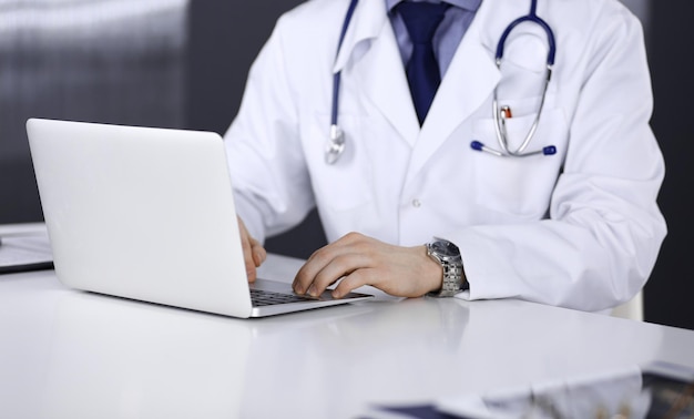 Unknown male doctor sitting and working with laptop in clinic at his working place, close-up. Young physician at work. Perfect medical service, medicine concept.