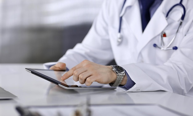 Unknown male doctor sitting and working with tablet computer in clinic at his working place, close-up. Young physician at work. Perfect medical service, medicine concept.