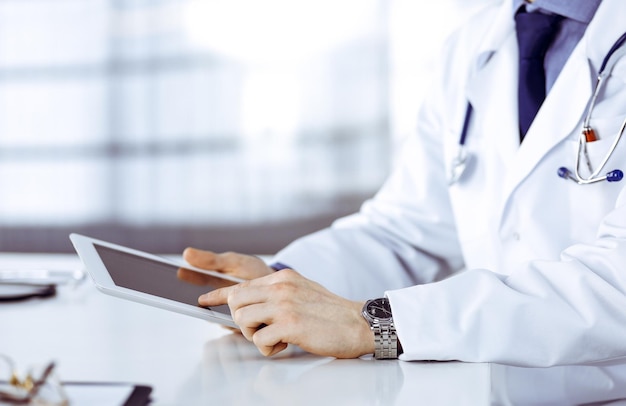 무명의 남성 의사가 자신의 직장에서 태블릿 컴퓨터로 앉아서 일하고 있습니다. 직장에서 젊은 의사. 완벽한 의료 서비스, 의학 개념입니다.