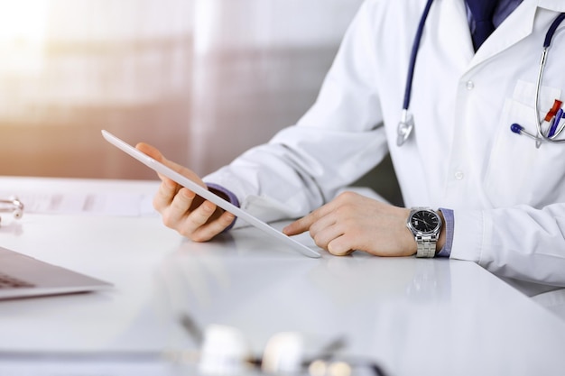 Unknown male doctor sitting and working with tablet computer iin a darkened clinic, glare of light on the background, close-up of hands