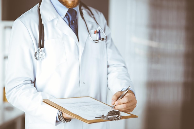 Unknown male doctor standing and writing with clipboard in clinic at his working place. Medicine concept.