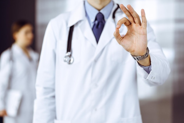 Unknown man-doctor standing straight and showing Ok sign in modern clinic. Female colleague is in a hurry at the background of physician. Medicine concept