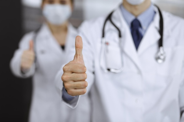 Unknown man-doctor and woman standing straight as a team and showing Ok sign with thumbs up in modern clinic. Medicine concept during Coronavirus pandemic. Covid 2019.