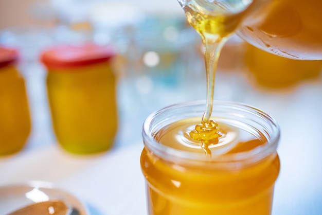 An unknown person pours sweet golden honey into transparent empty clean jars standing on a wooden white large table on a bright sunny day