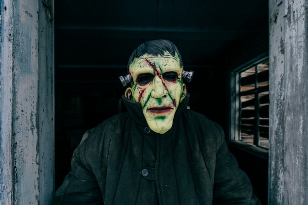 Photo unknown person with creepy horrible latex mask looking out from old wooden ghost house window. helloween concept.  spooky terrible fearful monster. children fear. frightening tales. nightmare.