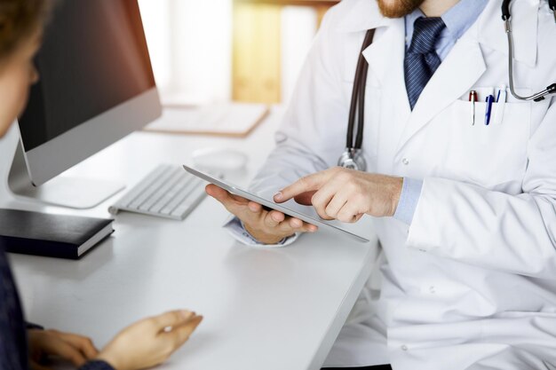 Unknown red-bearded doctor and patient-woman discussing current health examination while sitting and using tablet computer in sunny clinic, close-up. Medicine concept.