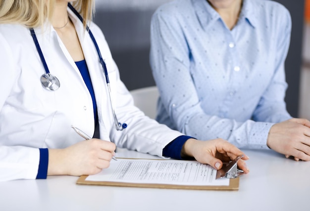 Unknown woman- doctor and her patient are looking at patient's current medical test results, while sitting together at the desk in the cabinet in a clinic. Female physician is using a clipboard and a