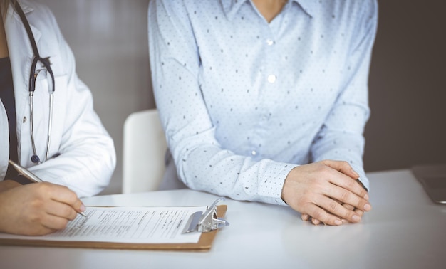 Unknown woman- doctor and her patient are looking at patient's current medical test results, while sitting together at the desk in the cabinet in a clinic. Female physician is using a clipboard and a