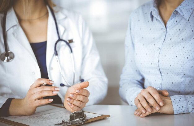 Unknown woman- doctor and her patient are looking at patient's current medical test results, while sitting together at the desk in the cabinet in a clinic. Female physician is using a clipboard and a