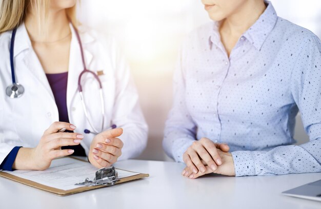 Unknown woman- doctor and her patient are looking at patient's current medical test results, while sitting together at the desk in the sunny cabinet in a clinic. Female physician is using a clipboard