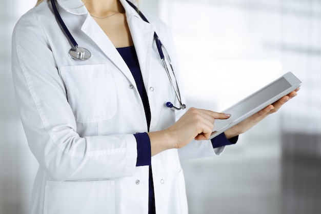 Unknown woman-doctor is holding a tablet computer in her hands, while standing in a clinic cabinet. Female physician at work, close-up. Perfect medical service in a hospital. Medicine concept.