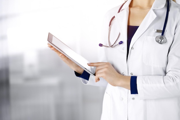 Unknown woman-doctor is holding a tablet computer in her hands, while standing in a clinic. Female physician with a stethoscope in her office, close-up. Perfect medical service in a hospital. Medicine