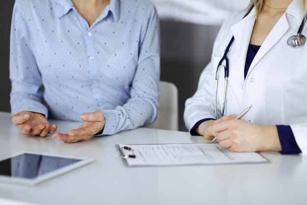 Unknown woman- doctor is listening to her patient, while sitting together at the desk in the cabinet in a clinic. Female physician with a stethoscope is writing at clipboard, close up. Perfect medical