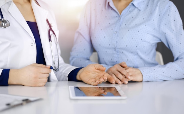 Unknown woman-doctor is showing to her patient a description of medication, while sitting together at the desk in the sunny cabinet in a clinic. Female physician is using a computer tablet and a steth