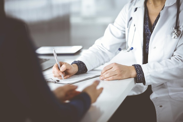 Unknown woman-doctor is writing some medical recommendations to her patient, while they are sitting together at the desk in the cabinet in a clinic. Physician is using a clipboard, close-up. Perfect m