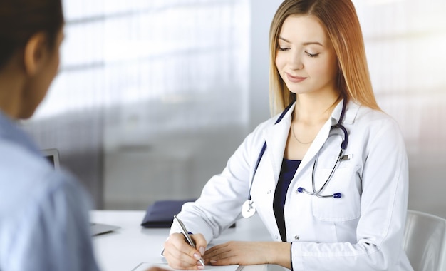 Unknown woman-doctor is writing some medical recommendations to her patient, while they are sitting together at the desk in the sunny cabinet in a clinic. Physician is using a clipboard, close-up. Per